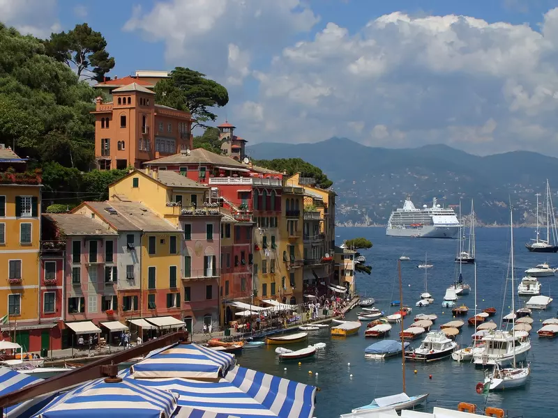 Colorful buildings line a boat-filled harbor with blue and white stripped umbrellas in the foreground