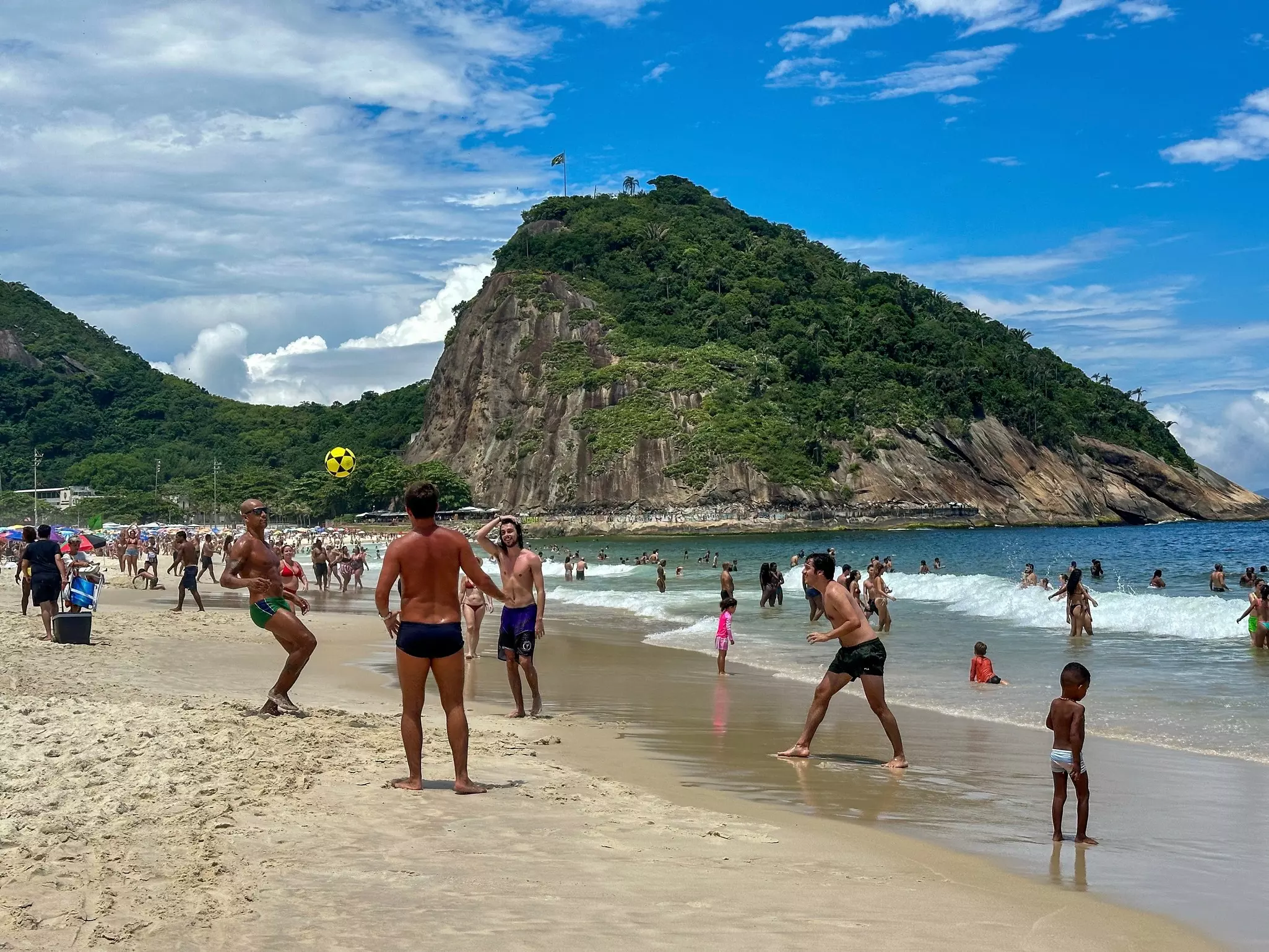 People in swimsuits kick a soccer ball on a city beach. A hill on a headland is seen in the distance.