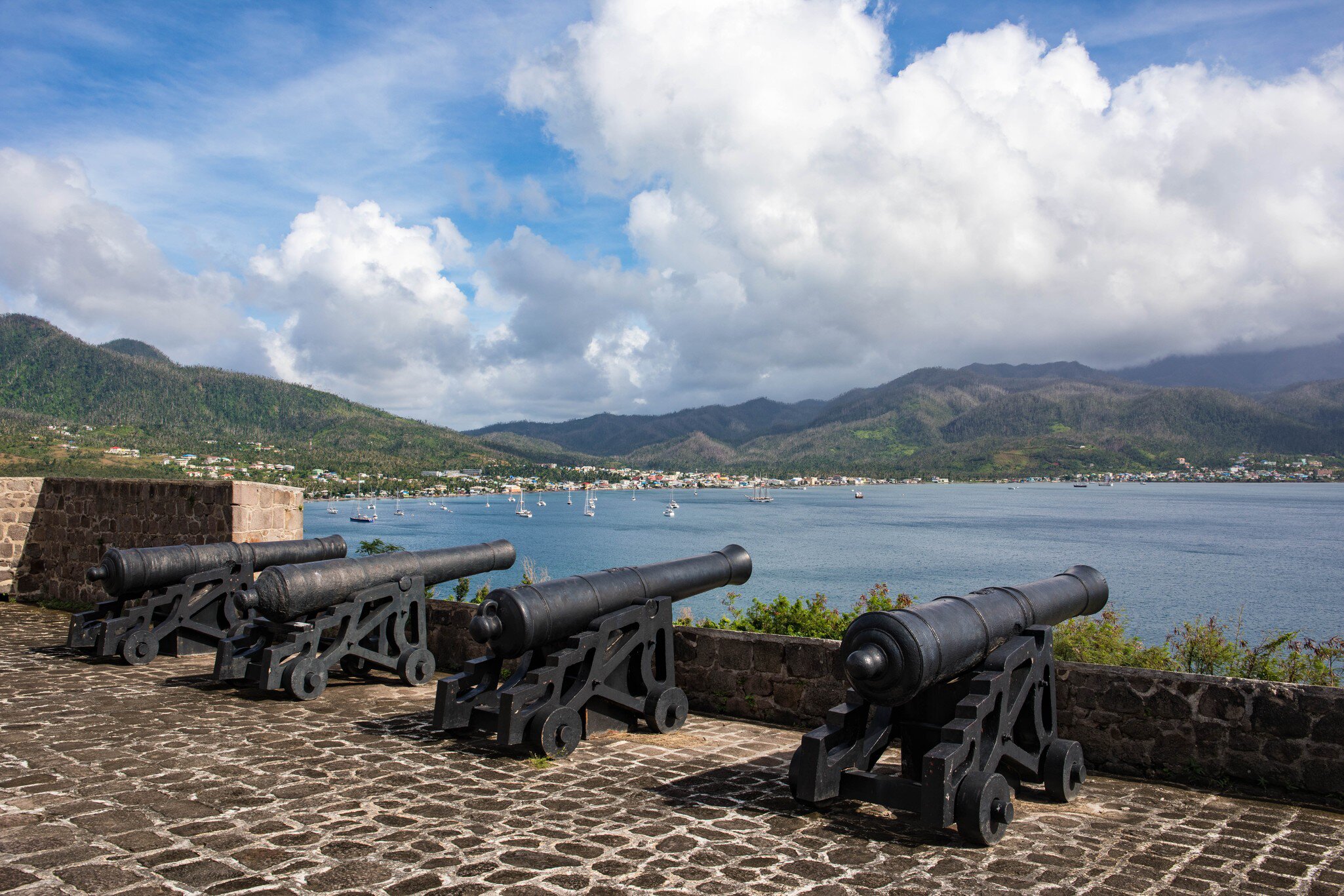 Four black iron cannon on wooden carriages along a cobbled battery above a bay of yachts, green mountains beyond.