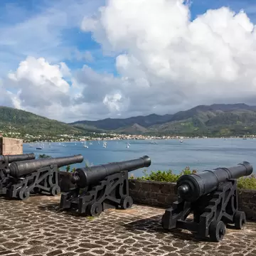 Four black iron cannon on wooden carriages along a cobbled battery, overlooking a bay of moored yachts with green mountains beyond.