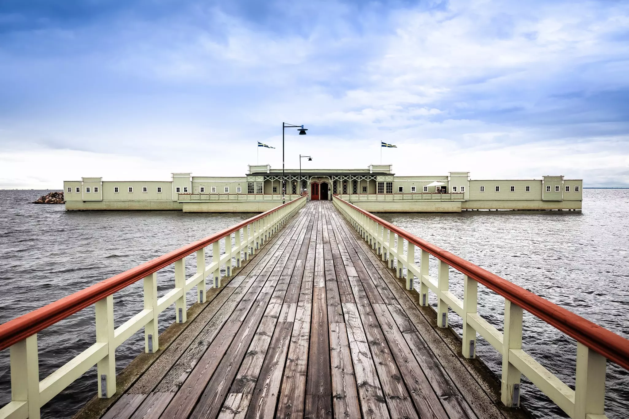 A low-slung, green-painted bathhouse on a pier is pictured, with a long walkway with wooden planks over the water leading to it.