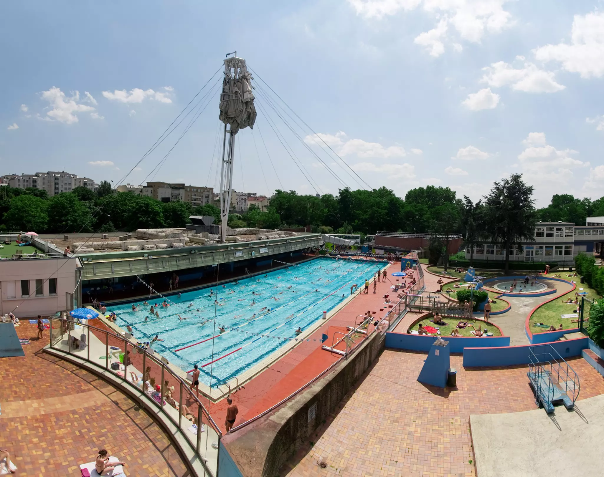 An Olympic-sized swimming pool on a sunny day. Swimmers use the lanes as people sunbathe on the grass nearby.