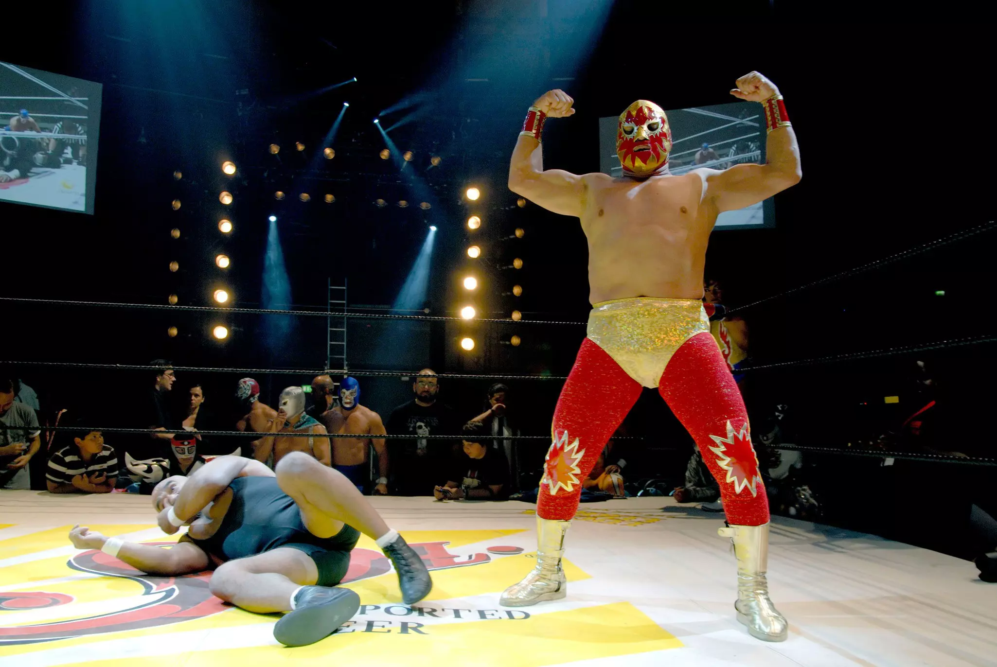 Lucha libre fighter in gold and red with a mask shows his muscles over a man in black lying on the mat. In the background people mill about and watch.