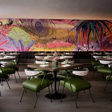 A room of dark tables set with glasses and flowers; the chairs have green seats and white backs, and there is a large colorful mural hanging.