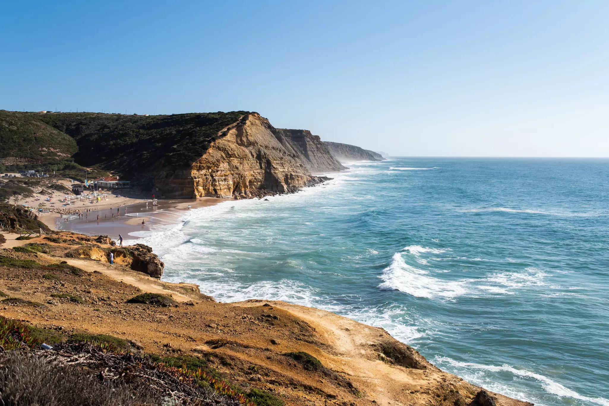 A small cove, surrounded by cliffs, is busy with tourists on a sunny day.
