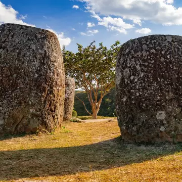 Two large weathered stone jars standing in dry grass beside a lone tree.