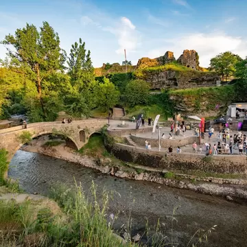 Stone arch bridge over the Ribnica River and a crowded riverside terrace below ruined fortress walls, Podgorica