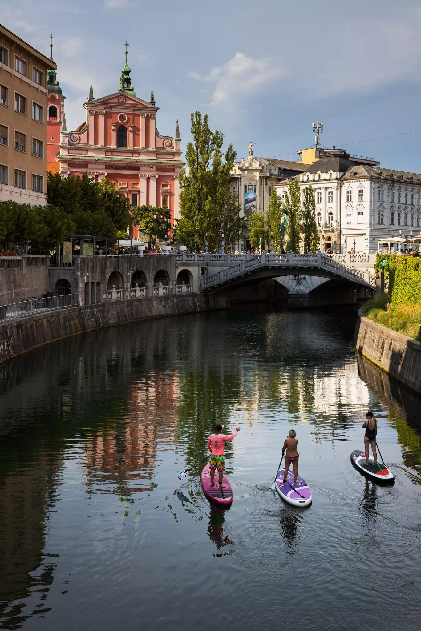 Three stand-up paddleboarders approach a bridge over a river in a city.