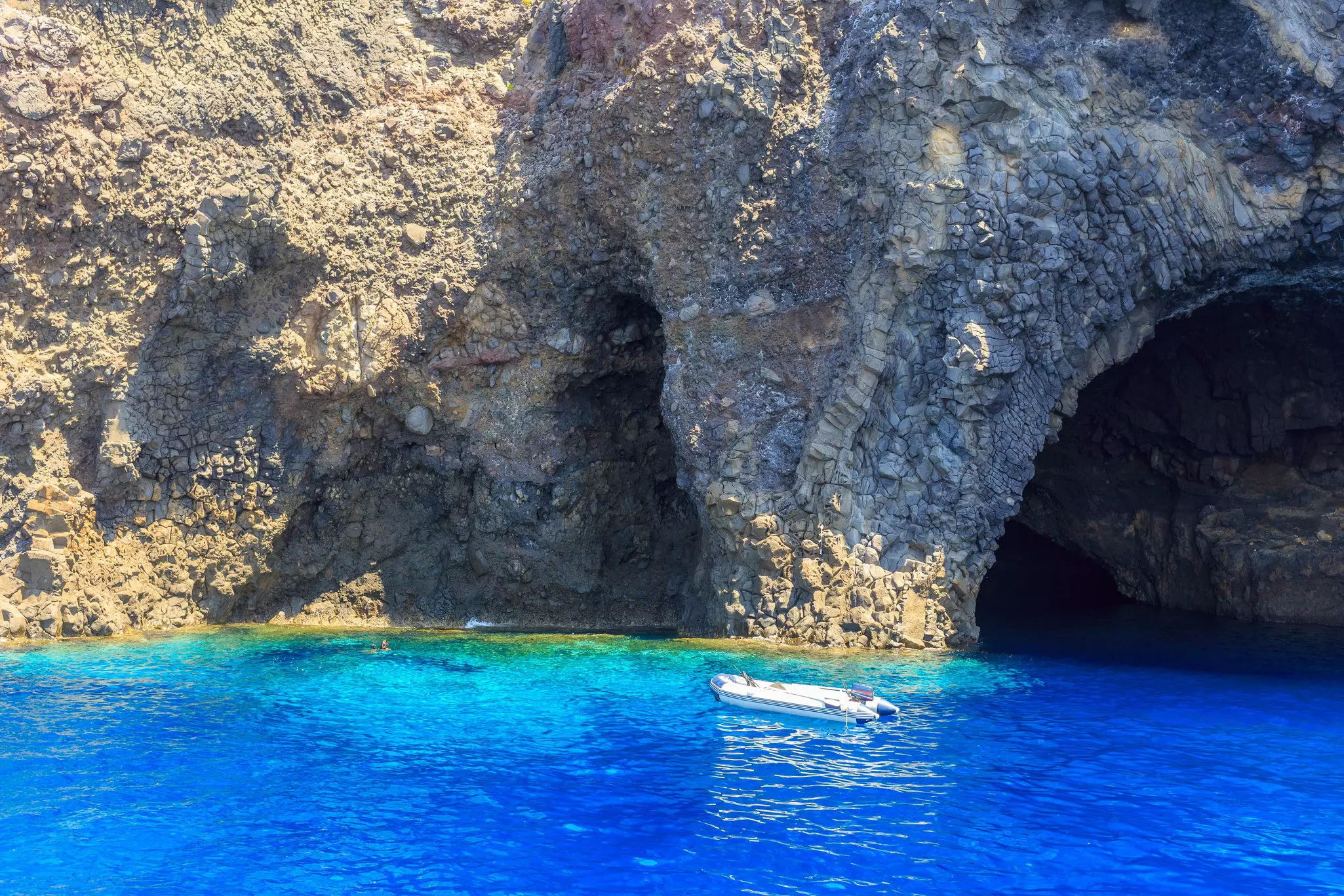 A small motorized boat moored in blue ocean near a cave formation. Two swimmers take a dip near the rocky coastline.