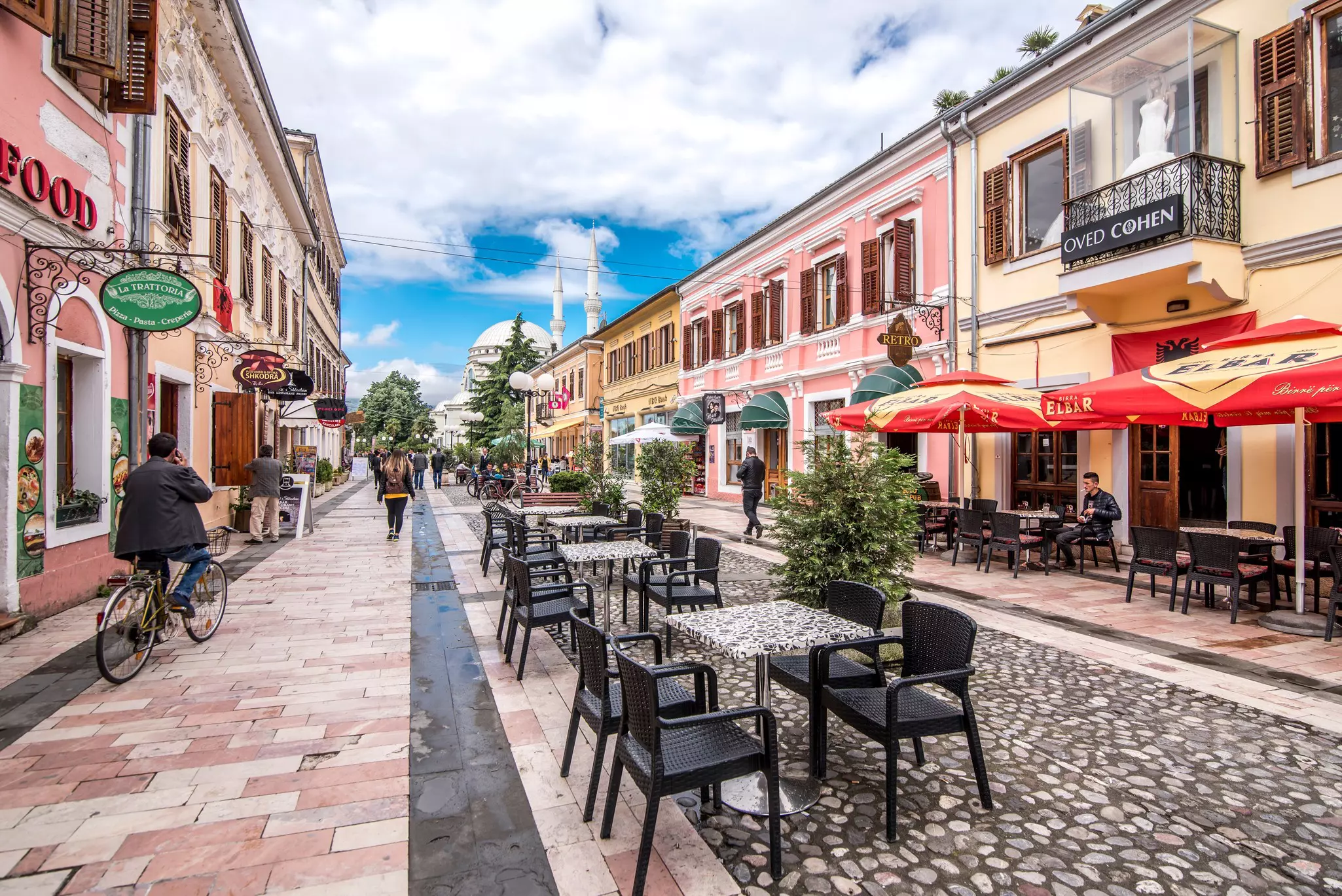 A cobbled street lined with pastel-colored buildings, home to bars, restaurants and shops. A white-domed mosque with two minarets is at the end of the street.