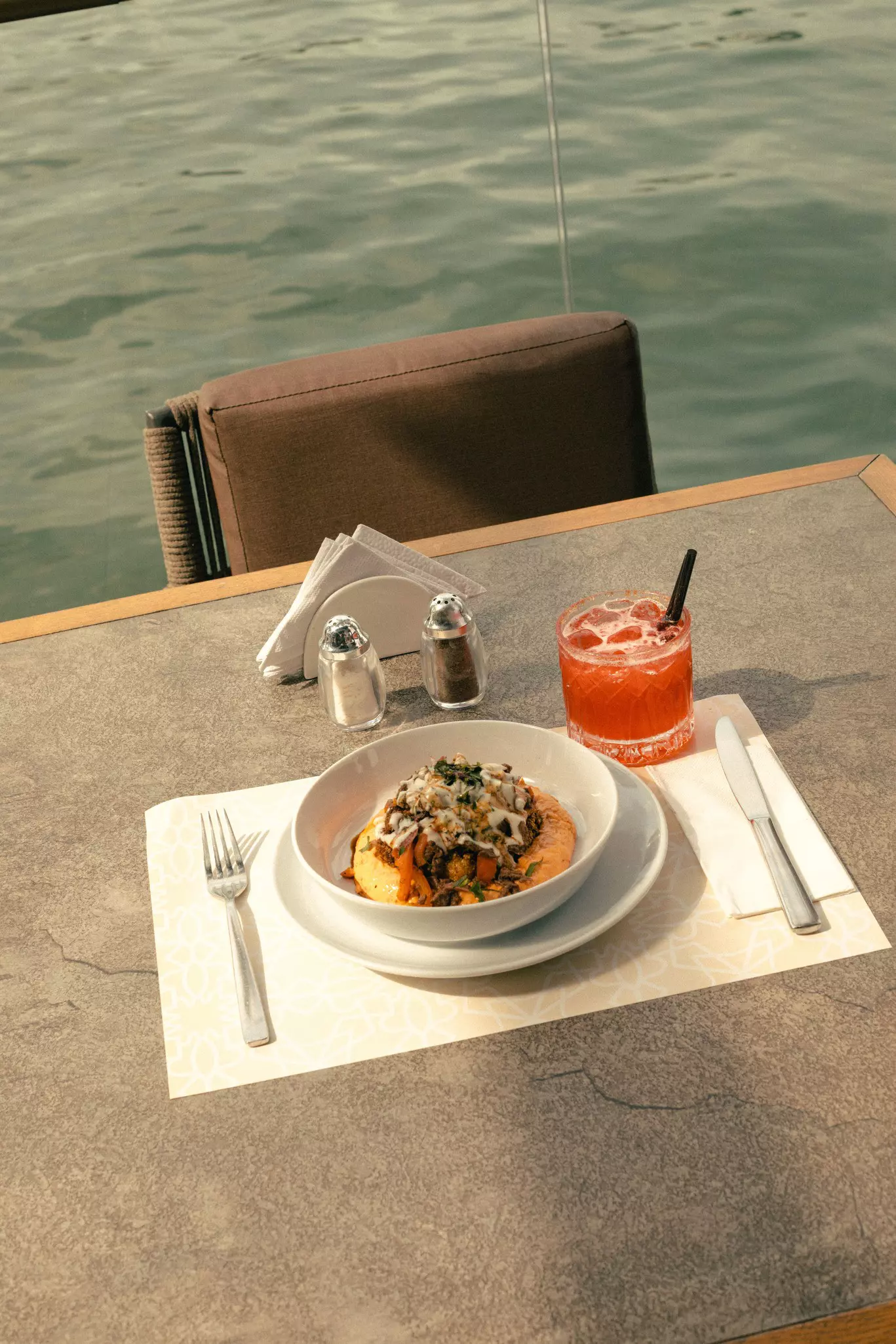 A bright coral cocktail and a bowl of Lebanese food on a table near the water