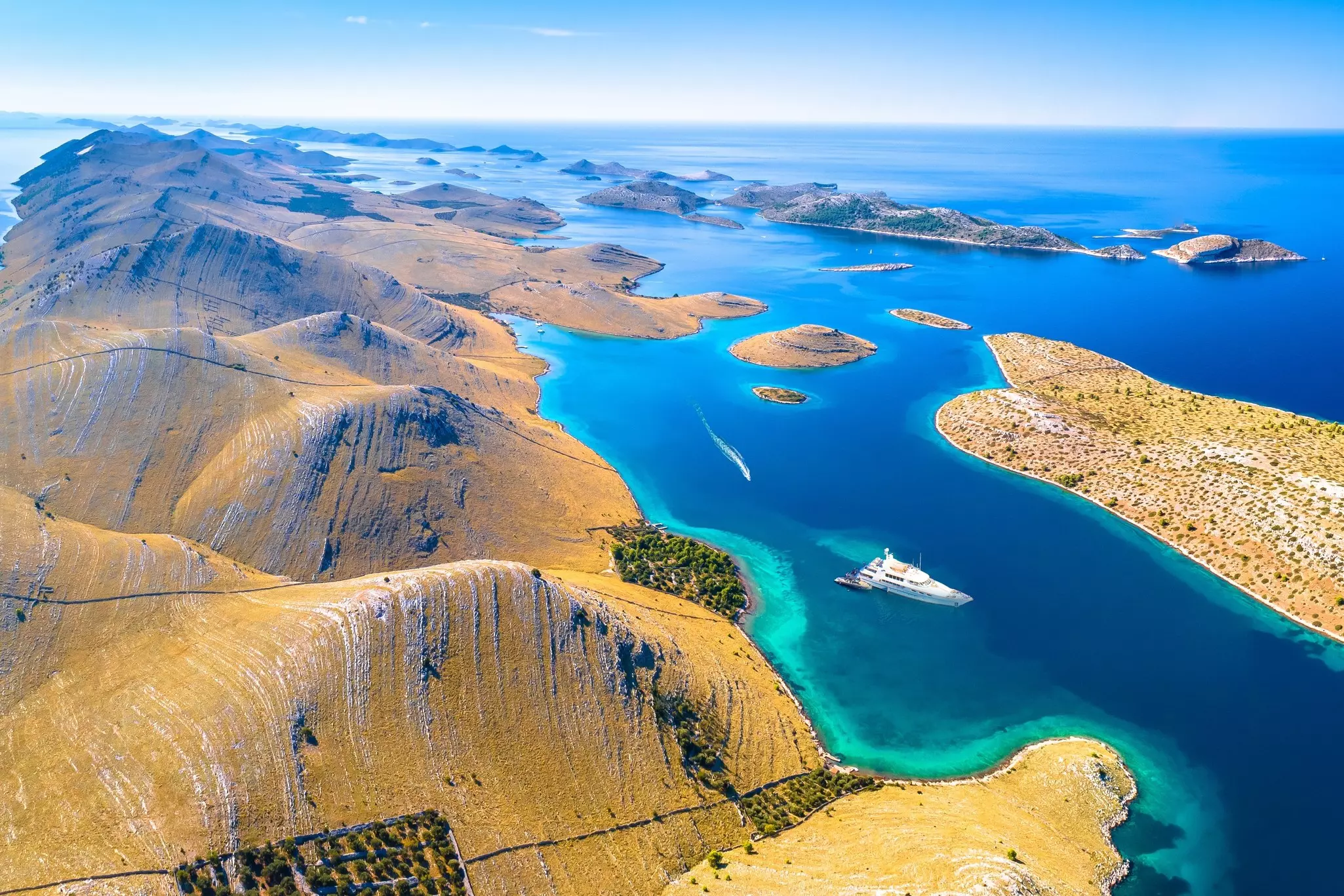 An aerial view of a yacht navigating the waters between hilly islands with scrubby vegetation.