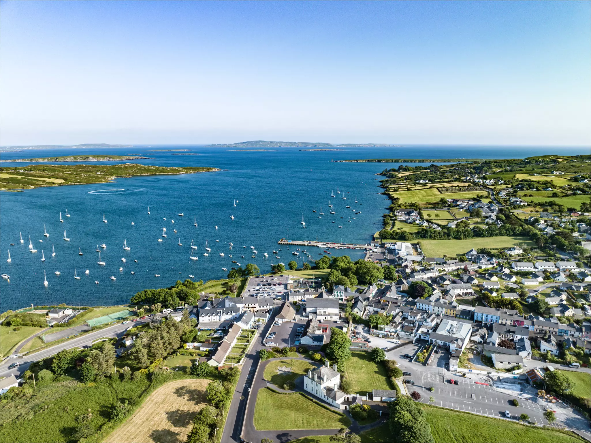 A harbor dotted with small sailing boats surrounded by green fields with a small village.