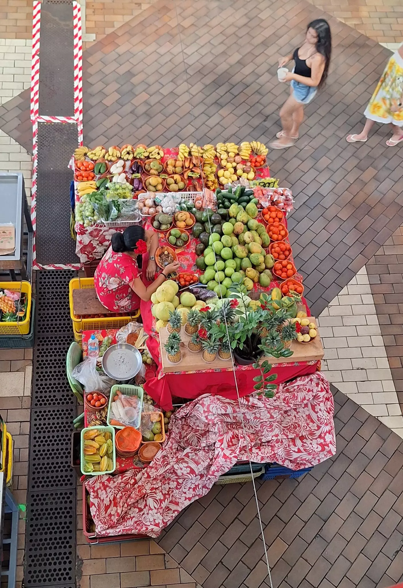 View from above of two people walking by a woman selling colorful fruits at an open-air market stall 