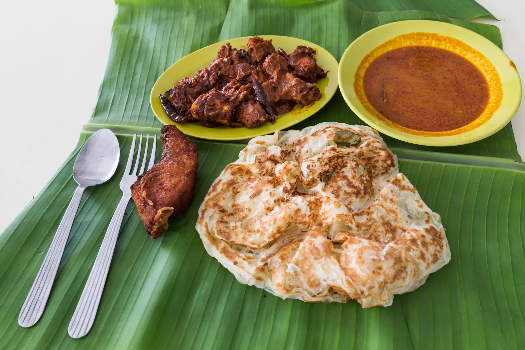 A flatbread is served on a banana leaf with meat and a bowl of curry.