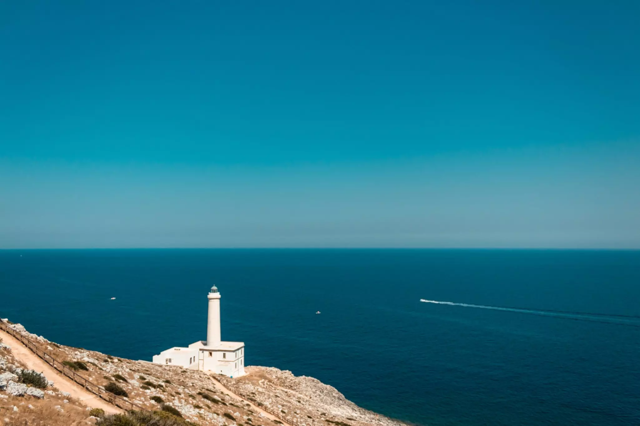 A white lighthouse is perched on a rocky cliff in Italy, with vast blue sea meeting the sky at the horizon.