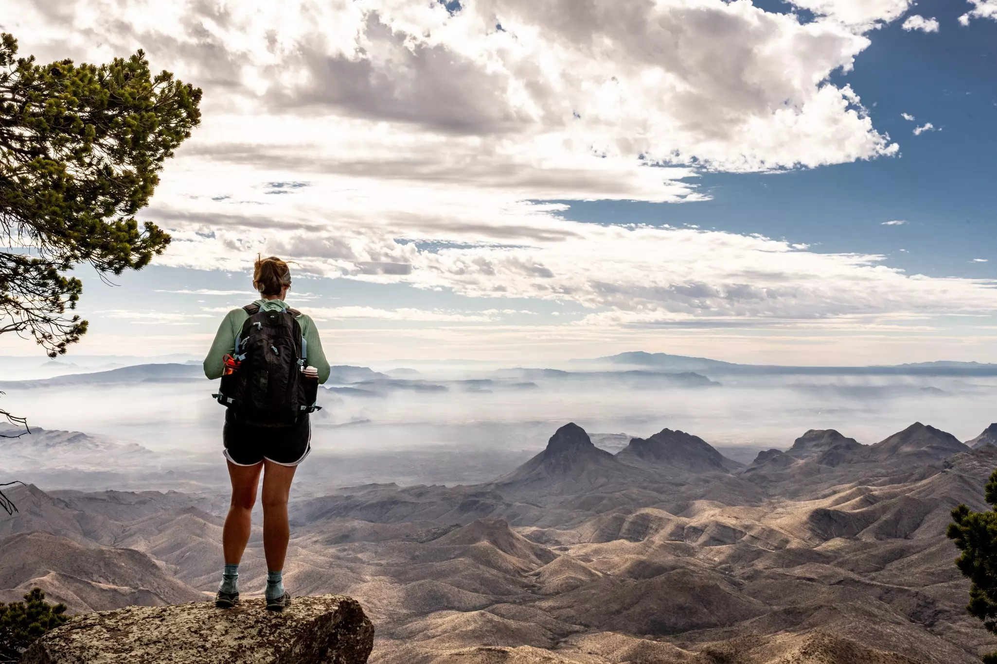 A hiker at the edge of a cliff looks down at a dry valley with low peaks; there are clouds on the horizon.