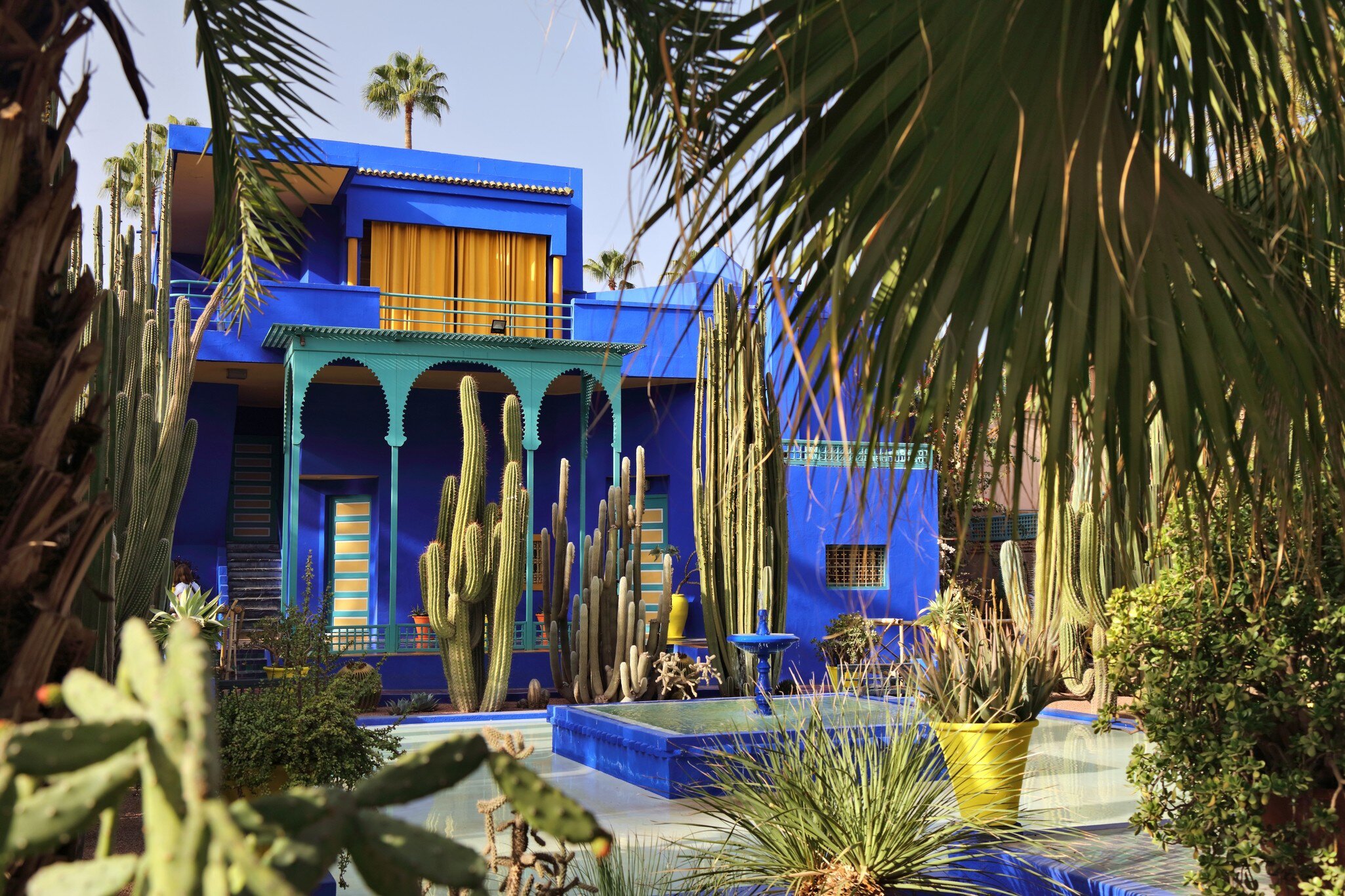 Cobalt-blue villa with a green arched loggia and yellow-striped doors behind tall cacti, palms and a blue pool.