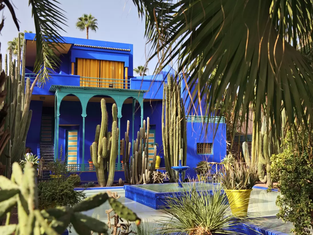 Cobalt-blue villa with a green arched loggia and yellow-striped doors behind tall cacti, palms and a blue pool.