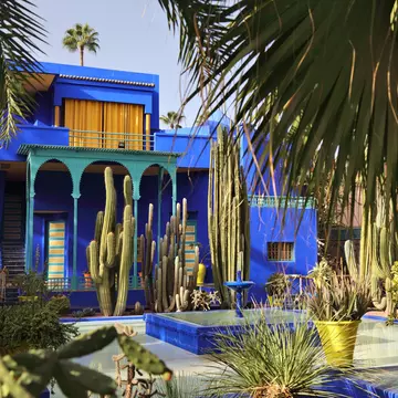 Cobalt-blue villa with a green arched loggia and yellow-striped doors behind tall cacti, palms and a blue pool.