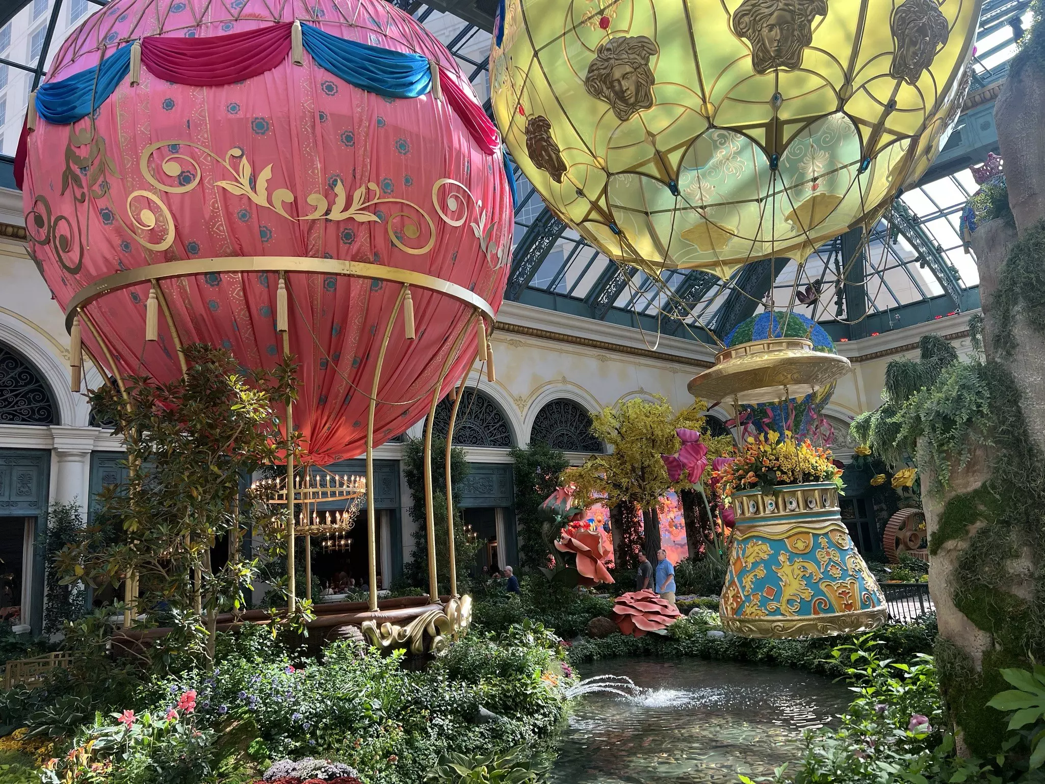 Outside view of the Garden Table (under the red hot-air balloon) located in the Bellagio Conservatory & Botanical Gardens. There's a second yellow hot-air ballon suspended above the garden. There are green plants, flowers and small pond in the foreground.