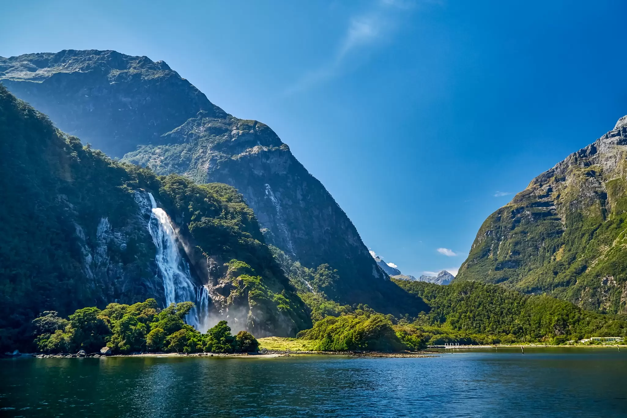 A tall waterfall cascading down a steep forested mountainside into the calm blue water of a fjord under clear skies