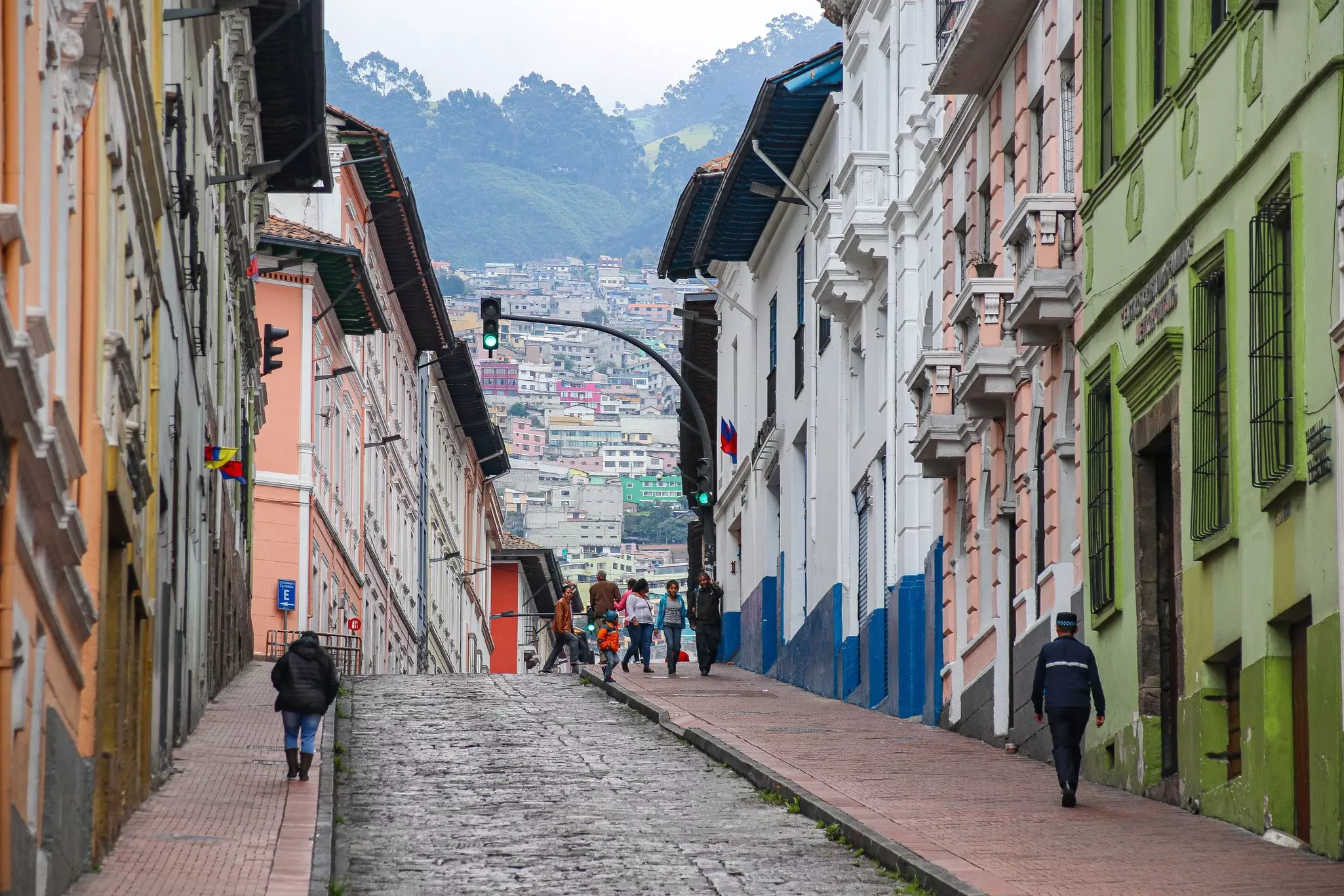 People walk up a steep city street fronted by historic buildings. Hillsides and mountains are visible in the distance. 