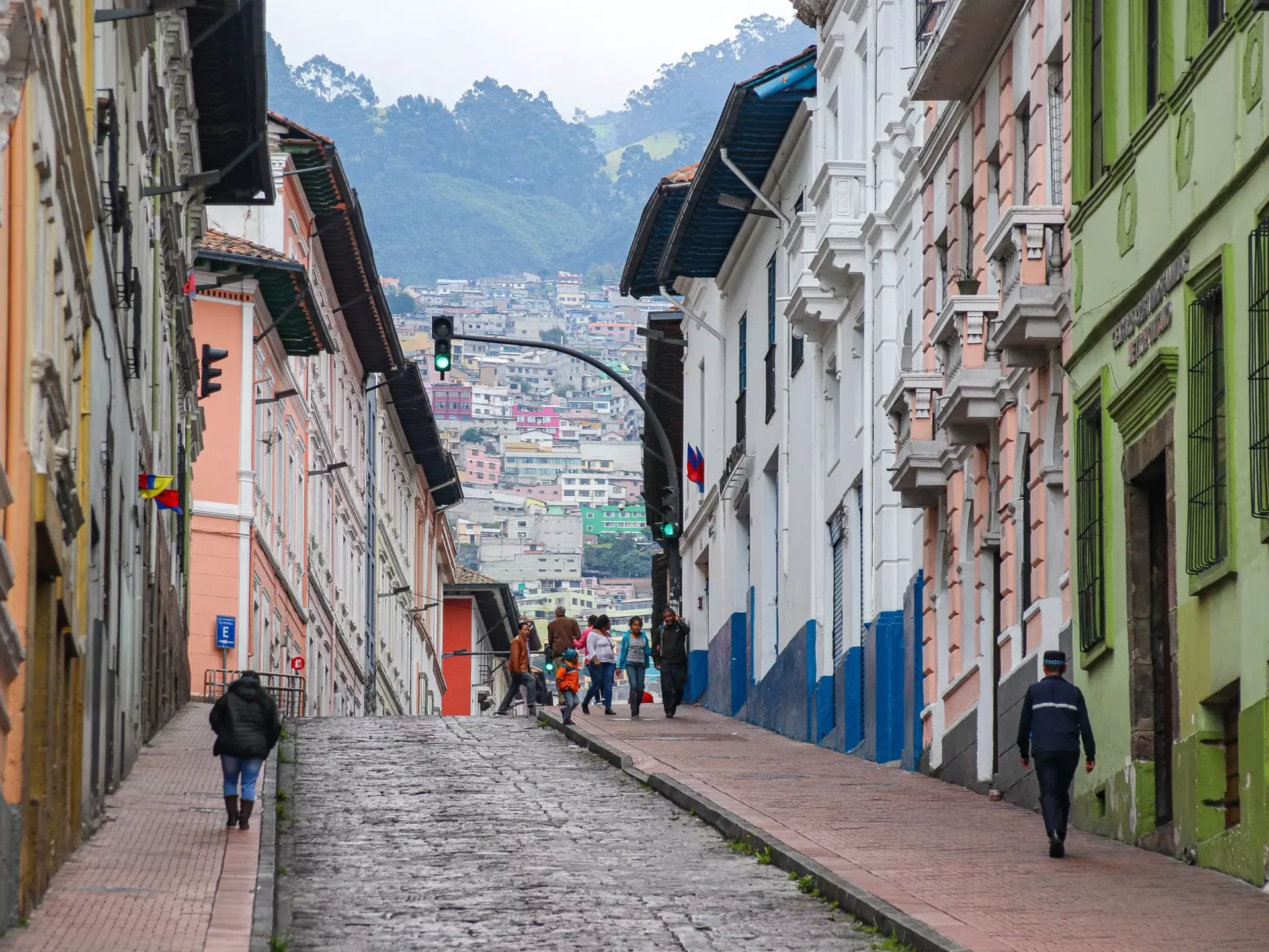 The scene on a city street in Quito in the historic center with locals walking and cars driving, set against a backdrop of hillside homes.