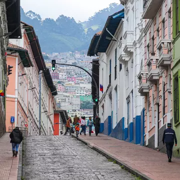 The scene on a city street in Quito in the historic center with locals walking and cars driving, set against a backdrop of hillside homes.