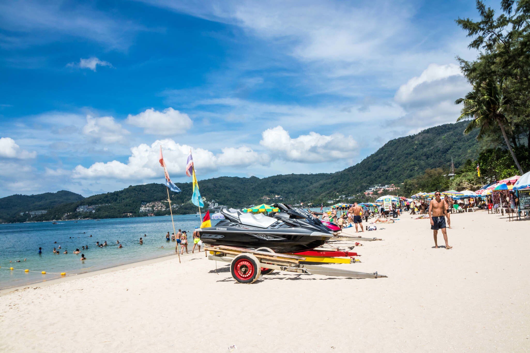 Jet skis on a trailer on a white sand beach with sunbathers, swimmers in the shallows and green hills behind, Patong, Phuket.