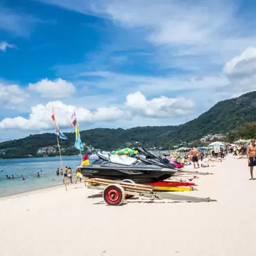Jet skis on a trailer on a white sand beach with sunbathers, swimmers in the shallows and green hills behind, Patong, Phuket.