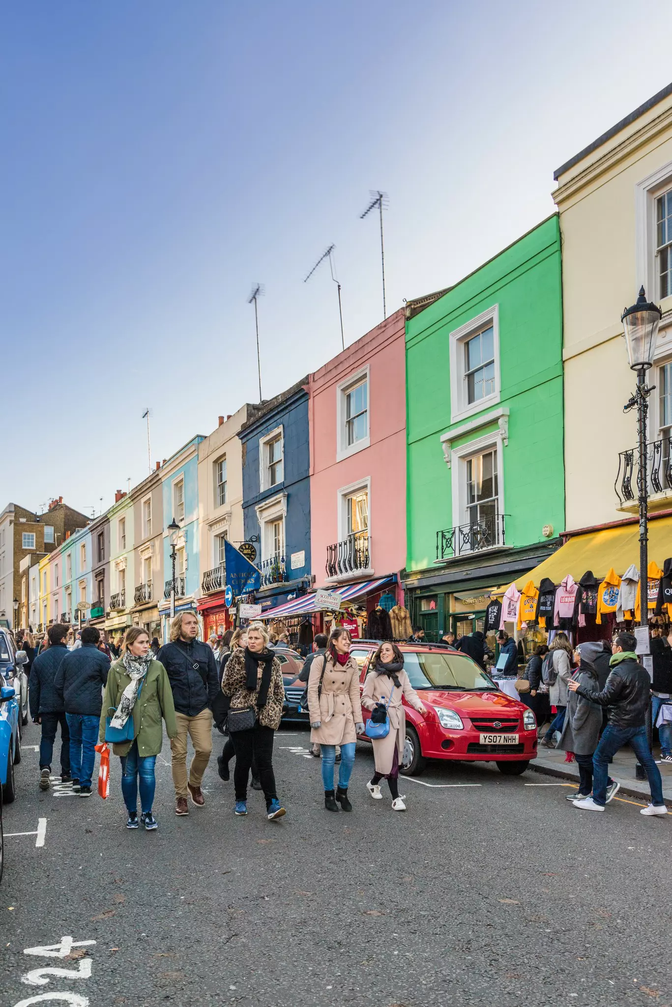 People wearing winter clothes walk down a street in front of a row of colorful houses, each with a market stall or shop below. 