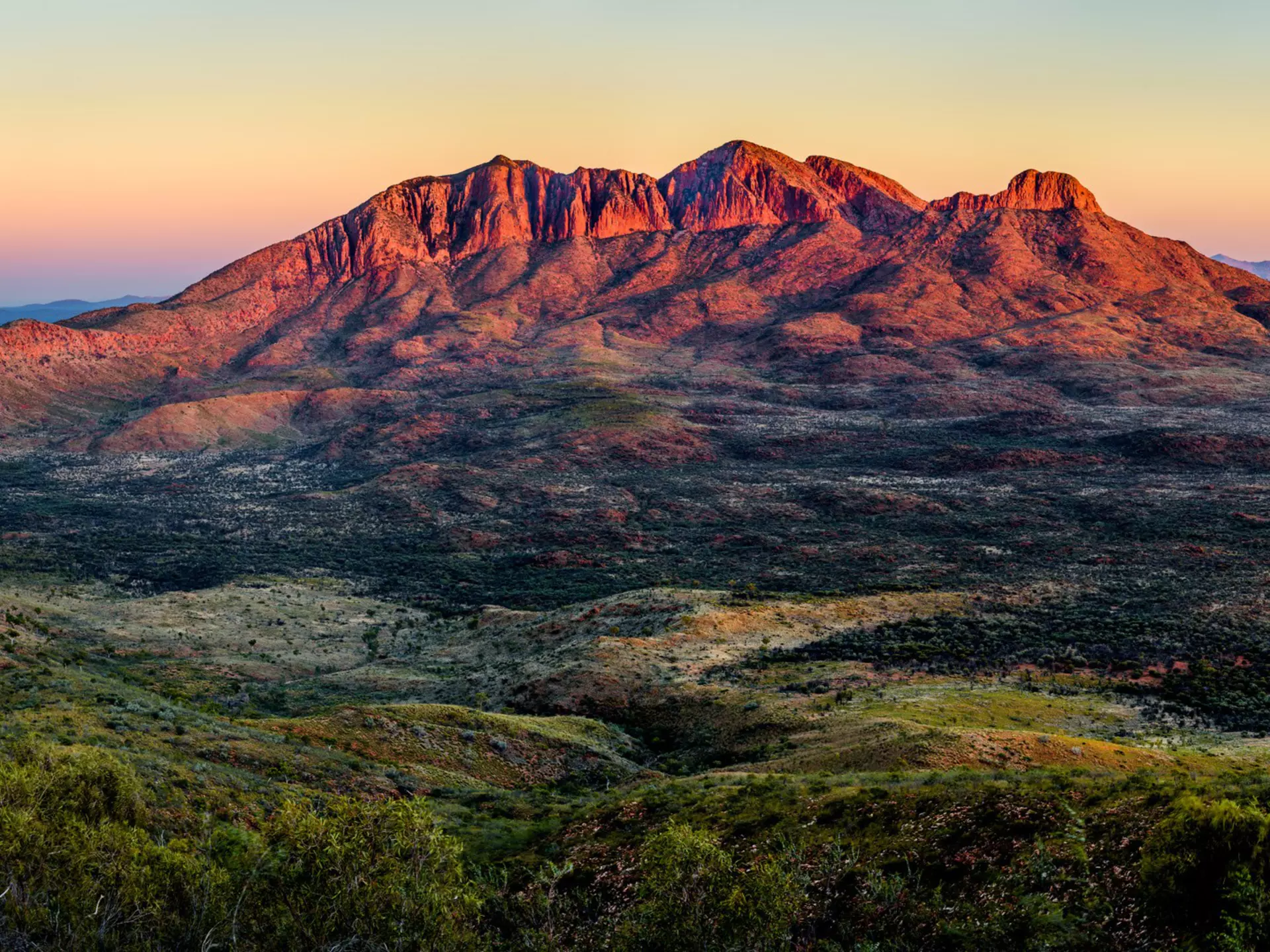 The deep red, orange, and purple alpenglow of Mt. Sonder in the West Macdonnel Range of Australia gleams at sunset over a variegated valley floor