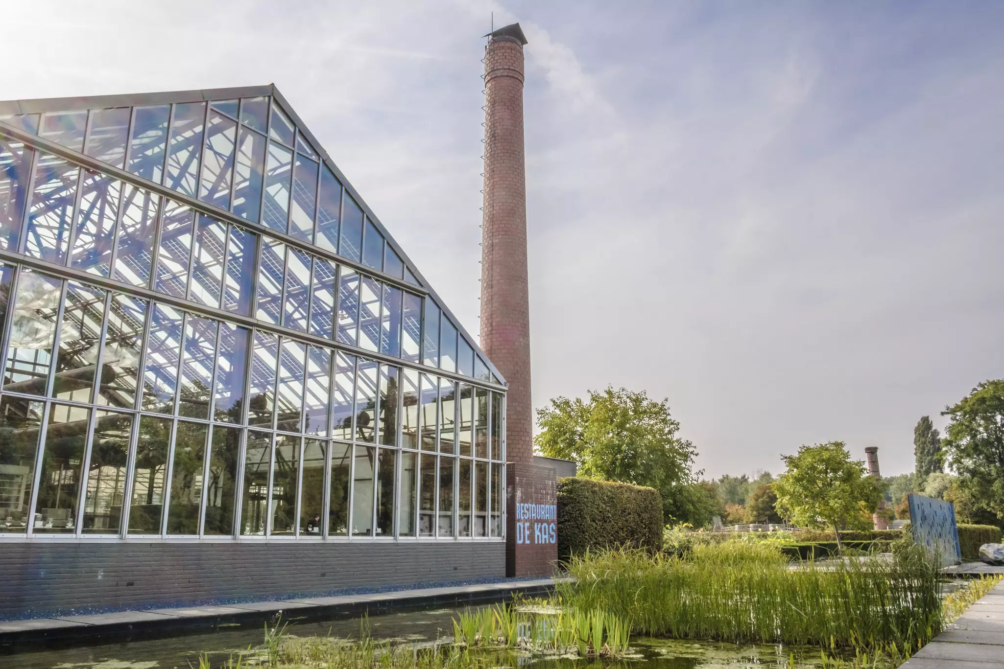 A large greenhouse restaurant with a red-brick chimney beside a pond