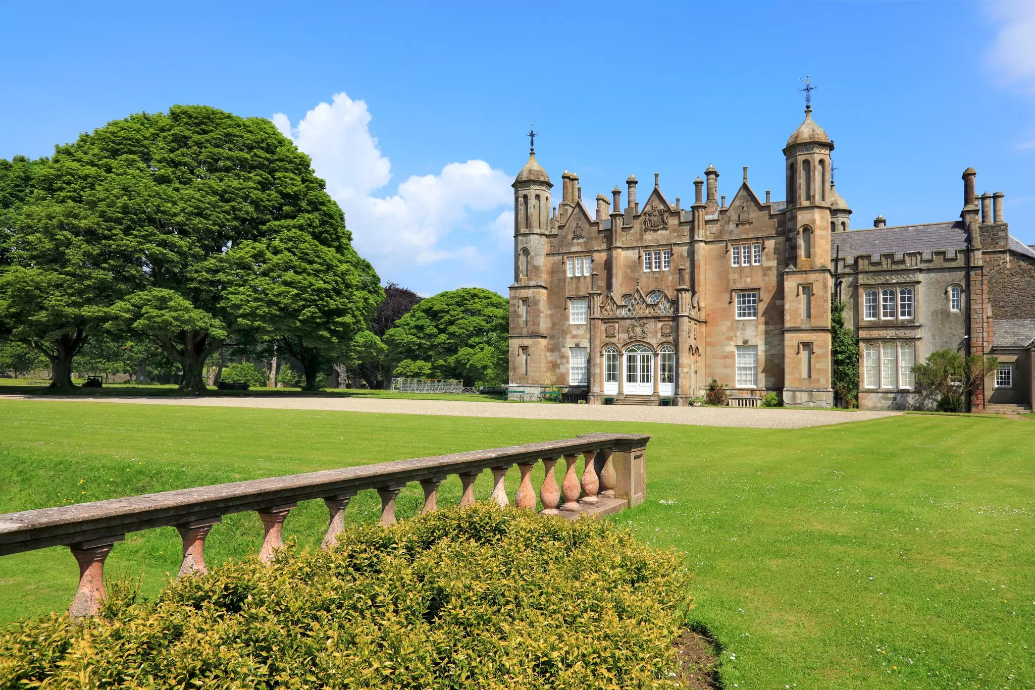 A stone balustrade is seen in a garden, with an ornate manor house across a well-mowed lawn.