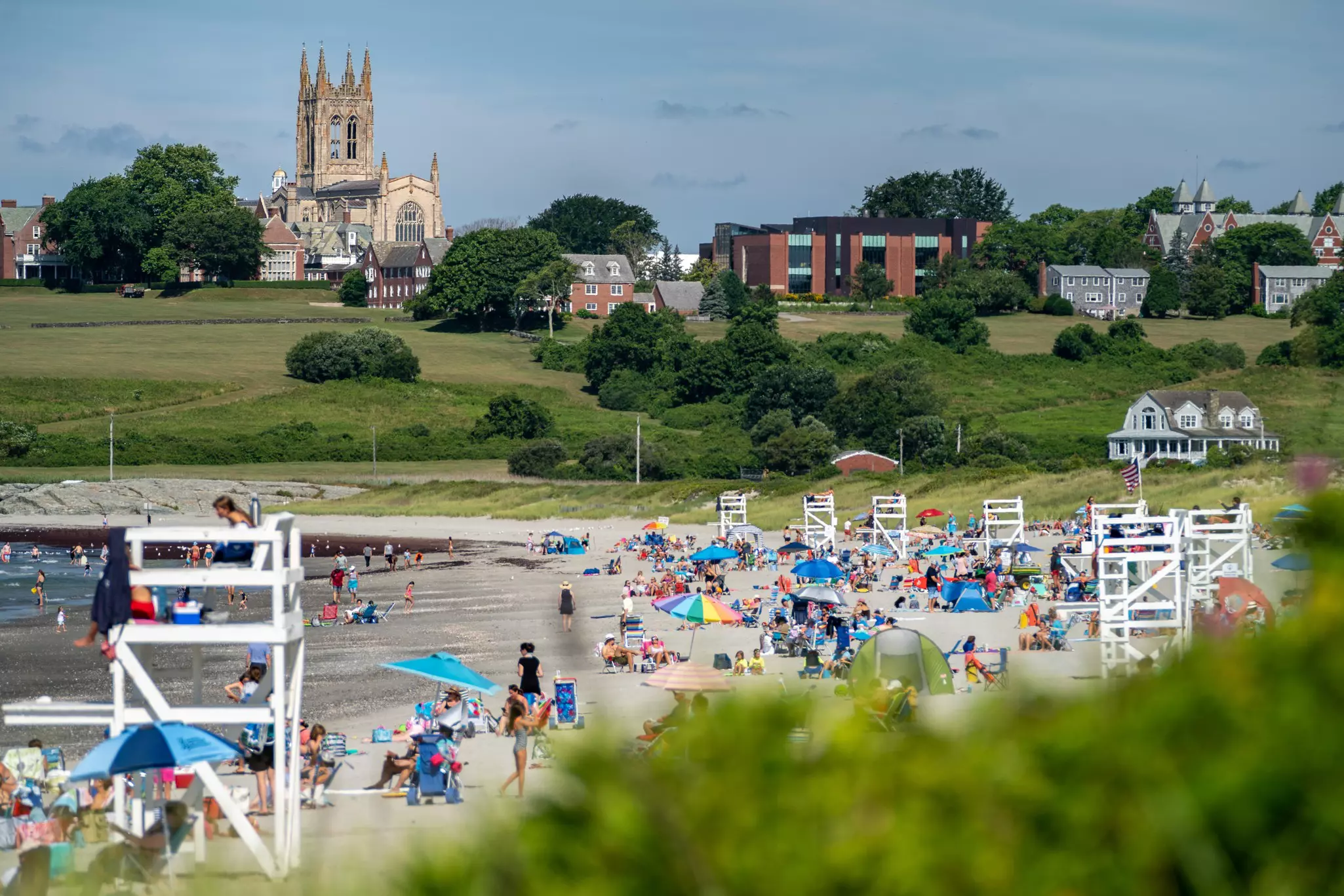 Sandy beach filed with beachgoers, umbrellas, and lifeguard stands, with a large gothic cathedral and brick buildings in the distance.