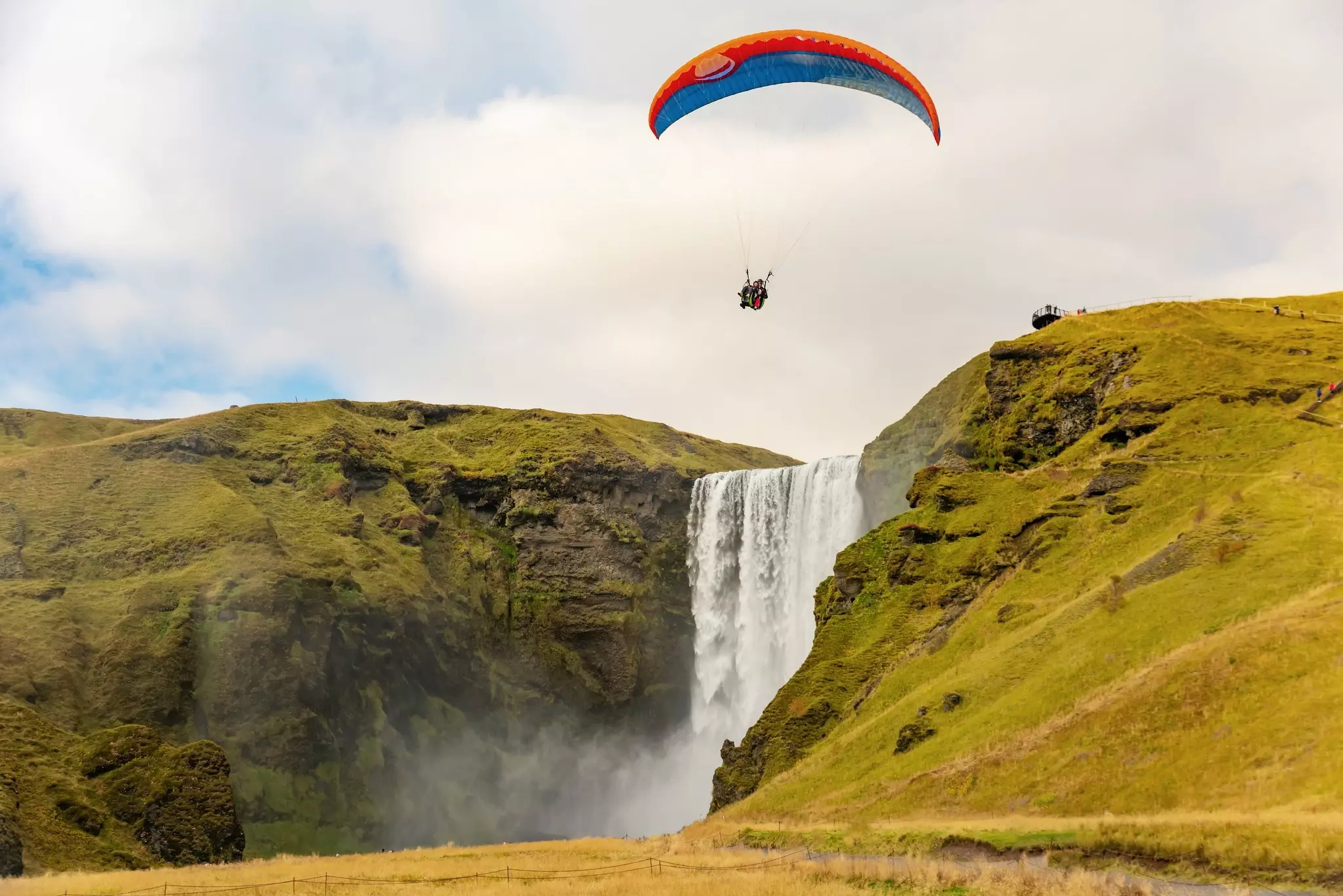 A tandem paraglider soars above cliffs covered with green vegetation and a roaring waterfall.