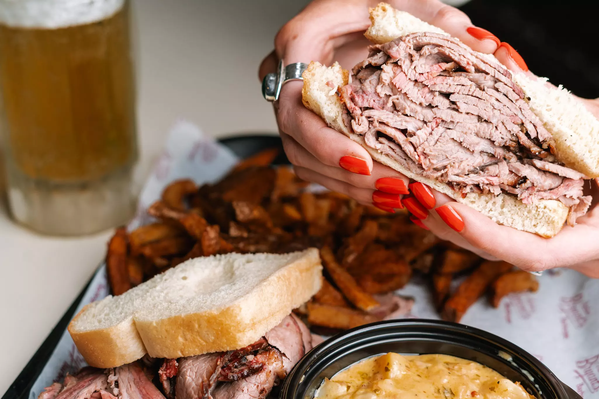 A close-up shot of woman with a red manicure holding a brisket sandwich over a plate of other food.