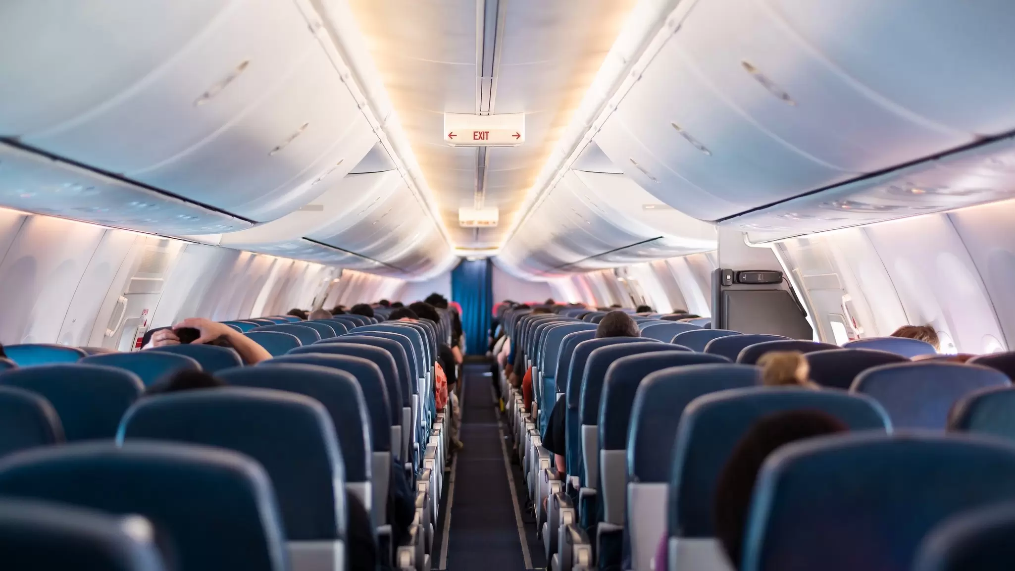 View from the back of an airplane aisle to the front, with overhead bag storage and rows of blue seats. 