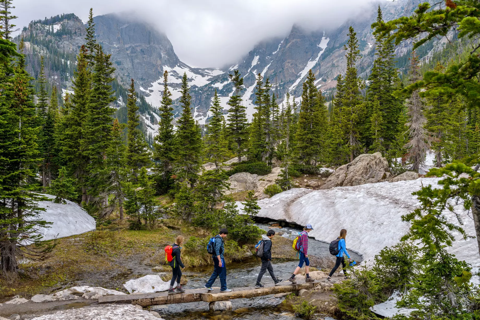 A group of hikers walking cross a tree trunk bridge over Tyndall Creek on Emerald Lake Trail in Rocky Mountain National Park, Colorado.