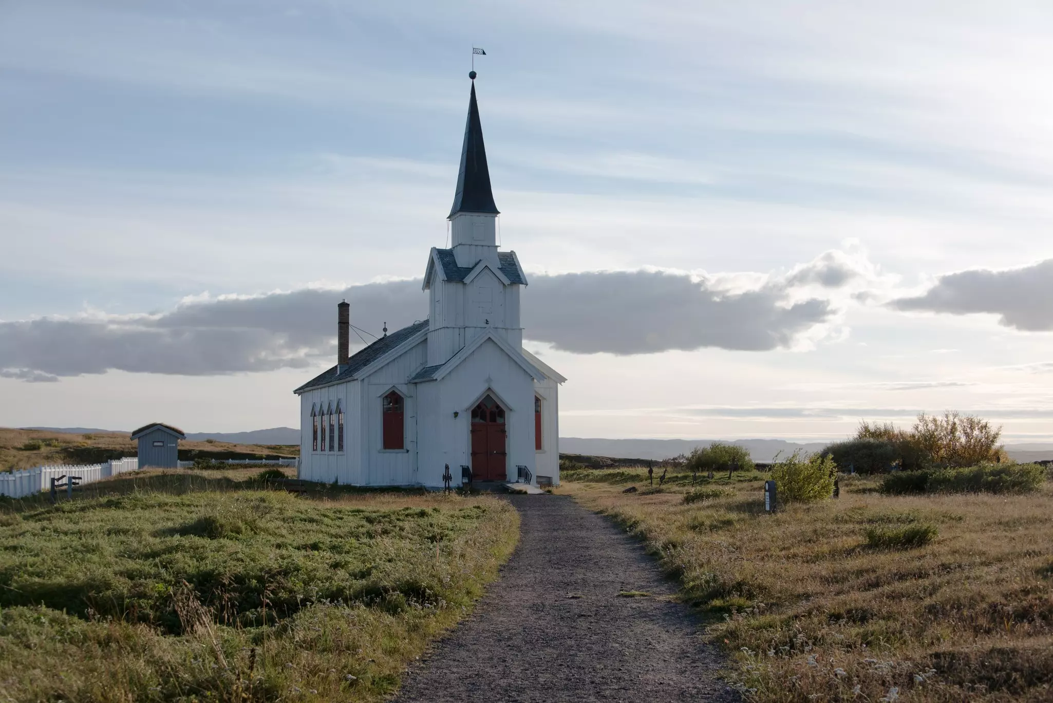 A small, plain, white church with spire at the end of a dirt path surrounded by low, autumnal grass on a partly sunny day.