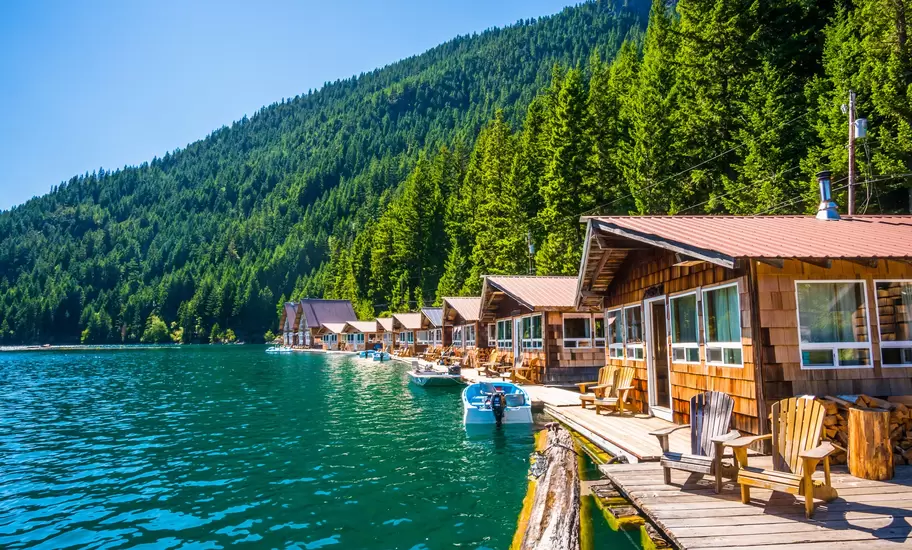 Floating wooden cabins with Adirondack chairs and boats on a turquoise lake below forested Washington Cascades slopes