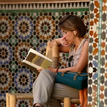 A woman sitting on a woven chair reading a book in a tiled courtyard.