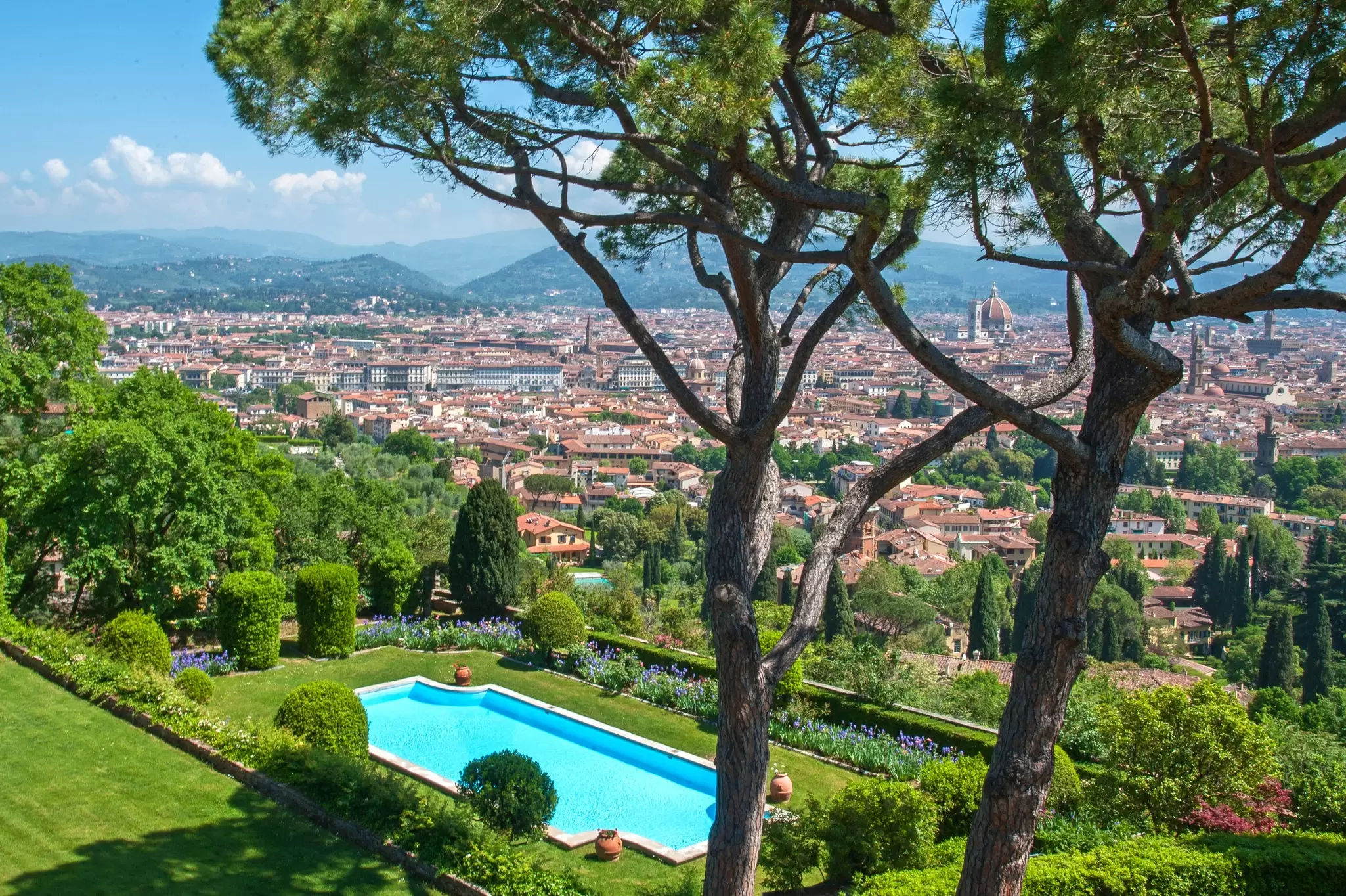 A luxurious hotel pool in a garden surrounded by trees, overlooking the skyline of Florence, Italy, covered in red-roofed buildings