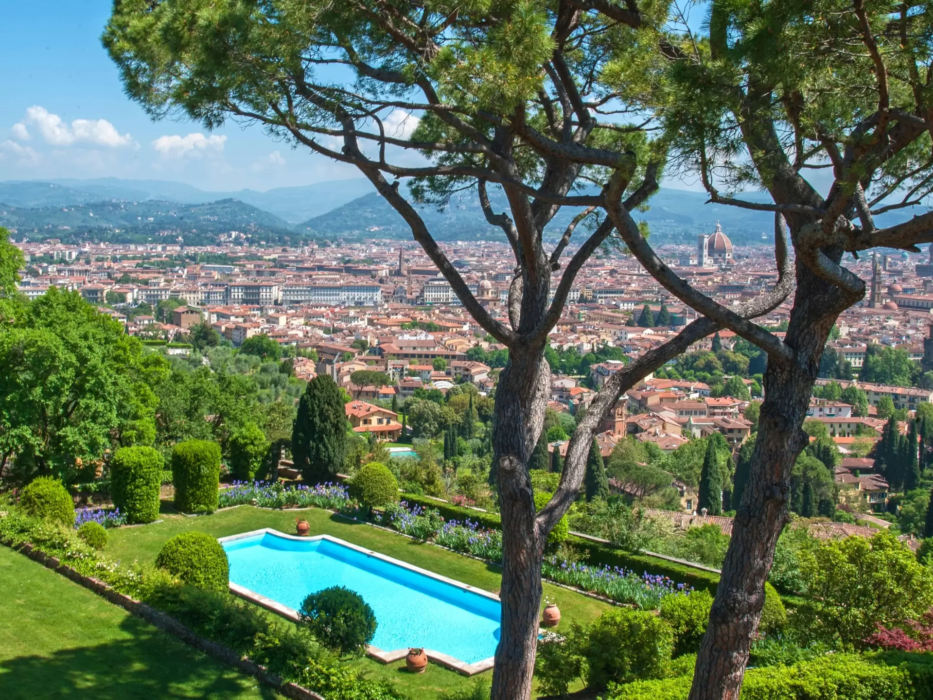 A luxurious hotel pool in a garden surrounded by trees, overlooking the skyline of Florence, Italy, covered in red-roofed buildings
