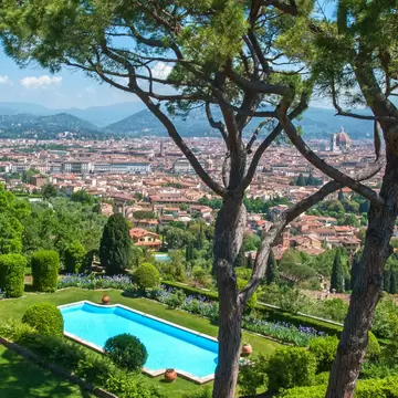 A luxurious hotel pool in a garden surrounded by trees, overlooking the skyline of Florence, Italy, covered in red-roofed buildings