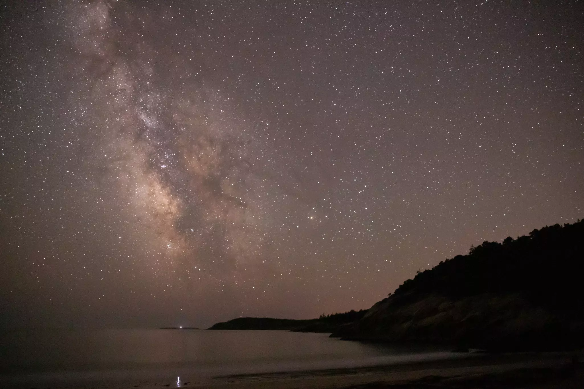 Dark night sky filled with stars and the brightness of the milky way galaxy above the ocean and a small mountain to the right.