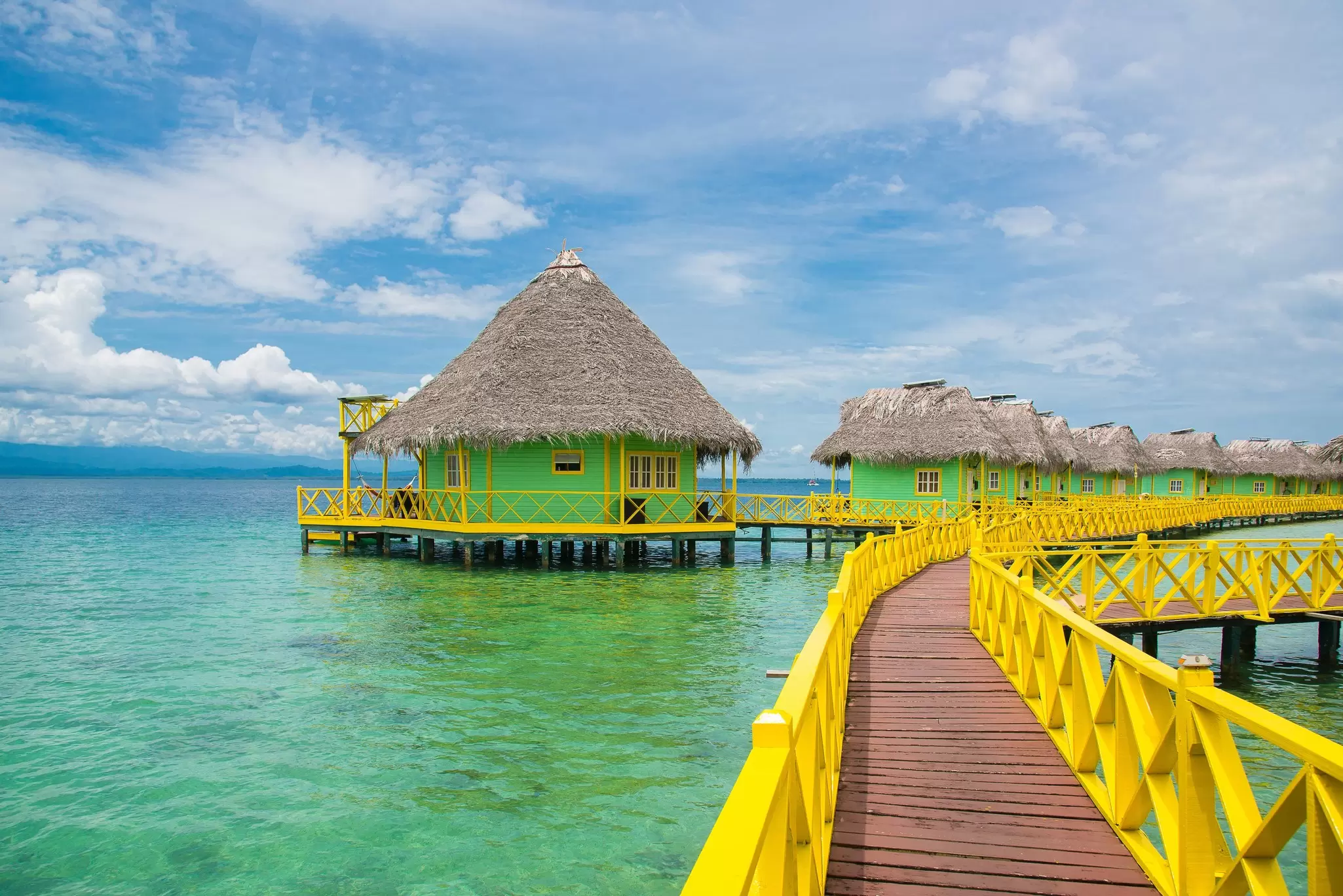 Wooden boardwalk with yellow railings leading to a series of green overwater villas
