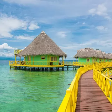 Wooden boardwalk with yellow railings leading to a series of green overwater villas