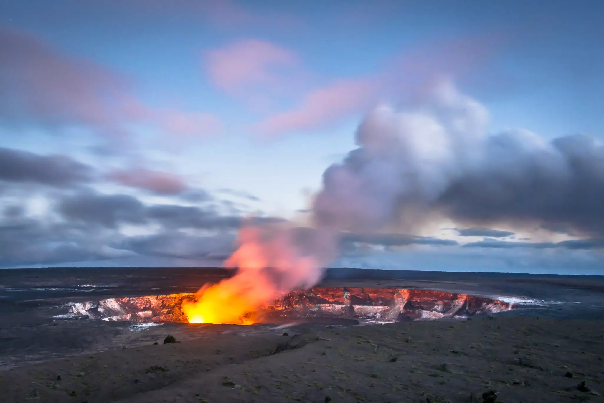 Plumes of smoke and steam rise from a volcano at twilight with the caldera glowing orange.